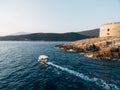 White motor boat with a canopy carries tourists along the coast in the background of ancient fortress. View from above