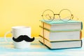 White mug with a felt moustache on the yellow background with stack of books and glasses. Concept of Father's day