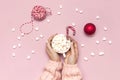 White mug with marshmallows in female hands in knitted sweater Candy Cane gifts boxes red ball on pink background top view