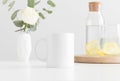 White mug mockup with flowers in a vase, glass and a bottle on a white table