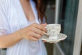 White mug with a pattern with hot coffee inside the in the hands of women in Bathrobe standing on the balcony of her bedroom. Woma