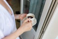 White mug with a pattern with hot coffee inside the in the hands of women in Bathrobe standing on the balcony of her bedroom. Woma