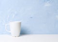 White mug with tea or coffee on a white table opposite a light blue concrete wall. Mocap with empty space for text.