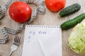 White notebook with pen and the inscription `my diet`, measuring tape and vegetables on a wooden table. The concept of weight loss