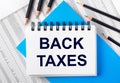 White notebook with the text BACK TAXES on the table next to black pencils on a blue background and reports. Business concept Royalty Free Stock Photo