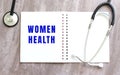 A white notebook with the words WOMEN HEALTH and a stethoscope on a gray wooden table.