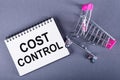 A white notepad with text COST CONTROL standing next to a shopping trolley on dark gray background Royalty Free Stock Photo