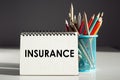 A white notepad with text Insurance standing next to a blue pencil holder on a white surface againsg black wall