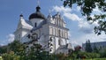 A white Orthodox church with domes, crosses, and a sheet metal roof. Surrounded by trees and flower beds. It is sunny