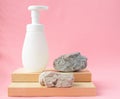 White packaging with a dispenser stands on a wooden stand and natural stones on a pink pastel background. Spa care concept, layout