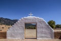 White painted mud adobe gate with cross on top looking out of a mission courtyard onto mountains and trees and a pueblo resident