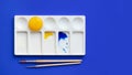 White palette with watercolor paints and brushes. Blue background. Instead of yellow, the egg yolk is used. Creative design and