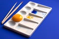 White palette with watercolor paints and brushes. Blue background. Instead of yellow, the egg yolk is used. Creative design and