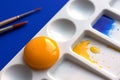 White palette with watercolor paints and brushes. Blue background. Instead of yellow, the egg yolk is used. Creative