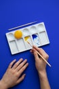 White palette with watercolor paints. Hands hold brushes. Blue background. Instead of yellow, egg yolk is used. Creative design
