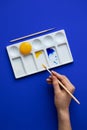 White palette with watercolor paints. Hands hold brushes. Blue background. Instead of yellow, egg yolk is used. Creative design