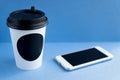 White paper kraft disposable cup for coffee with black plastic lid and white mobile phone on blue background Royalty Free Stock Photo