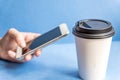 White paper kraft disposable cup for coffee with black plastic lid and white mobile phone in female hand on blue background Royalty Free Stock Photo