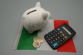 A white piggy bank with a calculator and euro coins over a flag of Italy. Italian financing concept for business and families