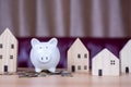 A white piggy bank is placed on a pile of coins. And next to it is a wooden model house. On the background is a red sofa. all on a Royalty Free Stock Photo