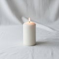 White pillar candle with a small flame sits on a smooth white fabric Royalty Free Stock Photo