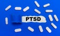 White pills lie on a beautiful blue background. In the center is white paper with the inscription PTSD. Medical concept. View from