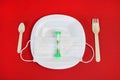 On a white plate, a medical mask and hourglass, wooden spoon and fork on a red background. Copy space - concept food, hunger,