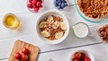 A glass of milk, juicy different berries, honey and a plate with granola and banana slices on a white wooden table