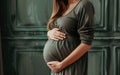 White pregnant woman holding hands on her belly. Long hair, feminine green dress, engagement ring, green wall, third trimester.