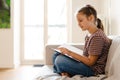 White preteen girl smiling and reading book while sitting on couch Royalty Free Stock Photo