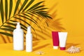 White pump bottles and colorful cosmetic tubes with no label next to shells and green palm twig on orange background