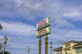 A white, red and blue sign at Johnnie Mercer\'s Fishing Pier with tall light posts, a building, lush green trees, blue sky