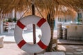 White red lifebuoy hang under tropical palm leaf umbrella, blurry hotel area background Royalty Free Stock Photo