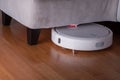 White Robot vacuum cleaner runs in corner under sofa on wood parquet floor. Modern smart cleaning technology housekeeping