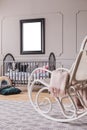 White rocking chair with pastel pink blanket in elegant little baby bedroom with grey wooden crib and mockup poster on the wall Royalty Free Stock Photo