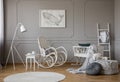 White rocking chair with pillow in the middle of cozy baby room interior with wooden cradle, industrial white lamp and poster in