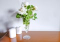 White roses in vase, white candles on wooden commode across white wall. Interior design. Home decor. Copy space