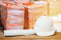 White safety helmet plastic, paper roll plan blueprint on wood floor table and pile brick orange background