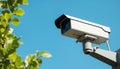White security camera mounted on pole against clear blue sky background. Green foliage visible on left. Modern surveillance tech Royalty Free Stock Photo