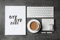 White sheet of paper with phrase Bye Bye 2020 near coffee, keyboard and wireless earphones on grey table, flat lay Royalty Free Stock Photo