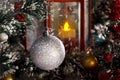 White shiny ball hanging on a branch of a Christmas tree against a red lantern with a candle