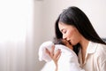 White shirt Asian mother is kissing at forehead of her newborn baby in bedroom in front of glass windows with white curtain to Royalty Free Stock Photo