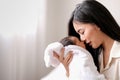 White shirt Asian mother is kissing her newborn baby in bedroom in front of glass windows with white curtain to show love and