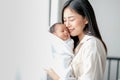 White shirt Asian mother is kissing her newborn baby in bedroom in front of glass windows with white curtain to show love and Royalty Free Stock Photo