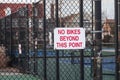 White sign with red lettering that says, No Bikes Beyond This Point on a metal chain link fence by a playground