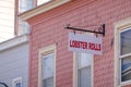 A white sign that says Lobster Rolls in red letters hanging from a house with pink wooden shingles