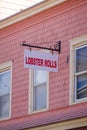 A white sign that says Lobster Rolls in red letters hanging from a house with pink wooden shingles