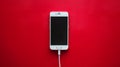 A white smartphone with a black screen connected to a white charging cable is positioned vertically on a bright red background. Royalty Free Stock Photo