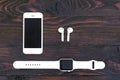White smartphone, smartwatch with a band and wireless bluetooth headphones on dark wooden background. View from above.
