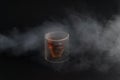 White smoke covers skull shaped glass of brown alcohol drink on black background.